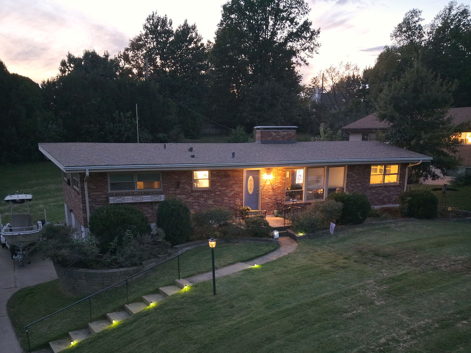 Aerial twilight view of a brick ranch home with lit windows, landscape lighting, and a long front lawn